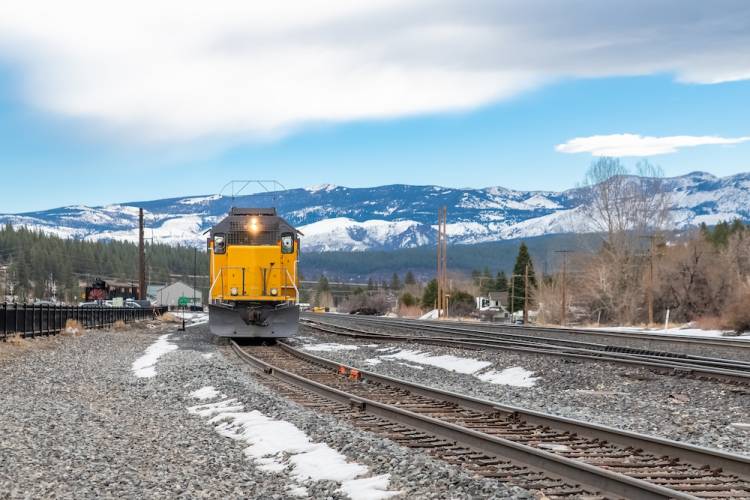 A yellow train on railroad tracks with snowy mountains in the background
